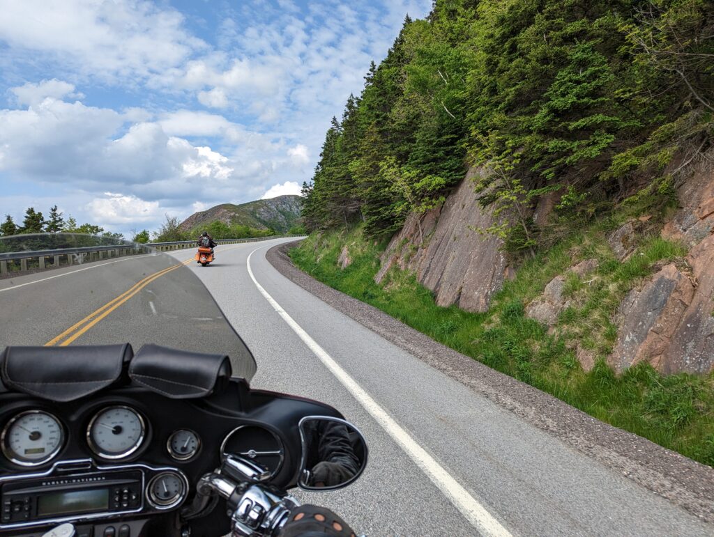 The perspective of Derek's motorcycle handlebars and Tam's Harley ahead of it about to negotiate a curve with rocks and trees on a steep cliff on the right. 