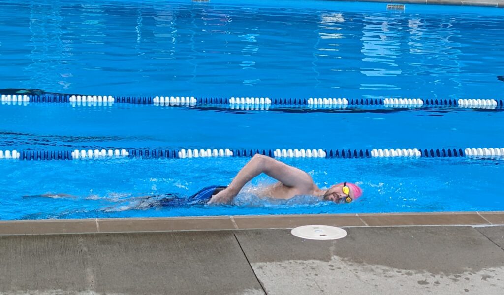 Jason swimming in the Paris pool. He's got a pink swimming cap, yellow goggles, and blue trunks on. 