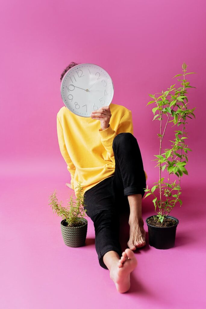 a person in yellow sweater sitting on the floor near the potted plants while covering face using a clock