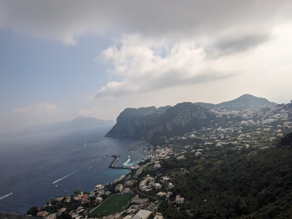A view of Capri from the top of the island. Rugged mountains are in view as well as houses, and lots of boats in the blue water. 
