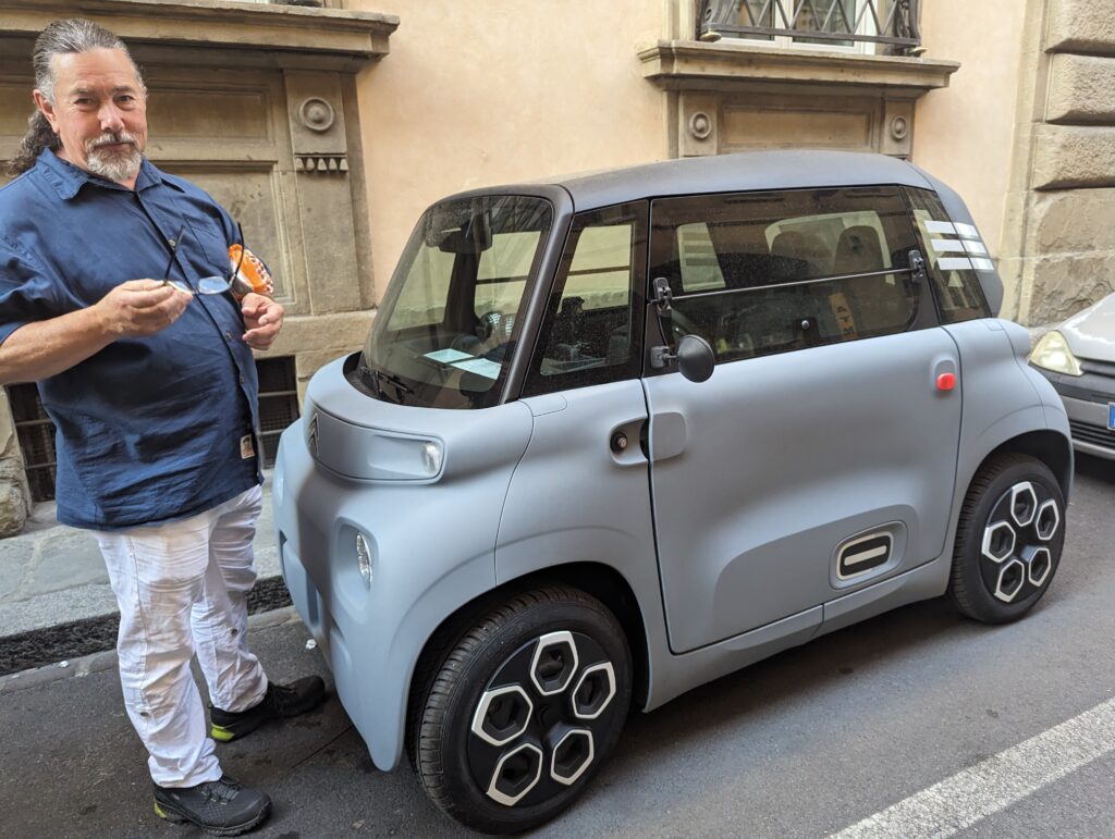 Derek stands beside a tiny, grey Citroen to demonstrate how tall it is. The top of the car is at his chest.