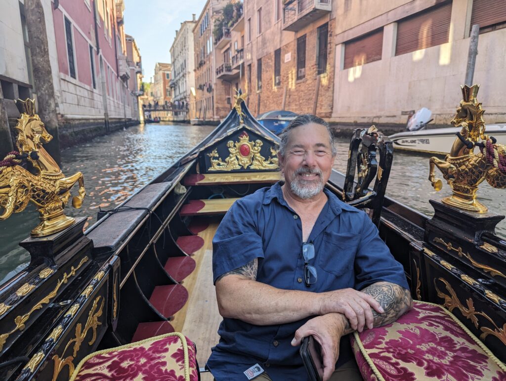 Derek grinning from ear to ear in our gondola ride. The gondola is shiny black and decorated ornately with gold-toned details and red brocade fabric on the seats. 