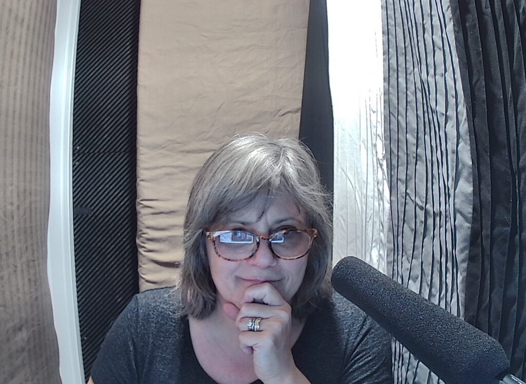 Me in my recording booth one year ago, looking toward but not at the camera, with my long, shotgun microphone in the forefront.