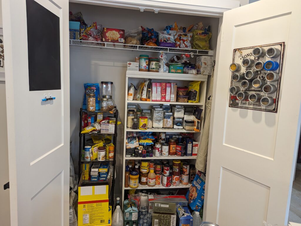 Cluttered pantry is the size of a big closet. It has canisters and packages all stuffed into a bookshelf and another, smaller shelf. Spices are in magnetized containers attached to a metal Farm Produce sign on the inside door. On the other door is a blackboard (painted on) with chalk. 