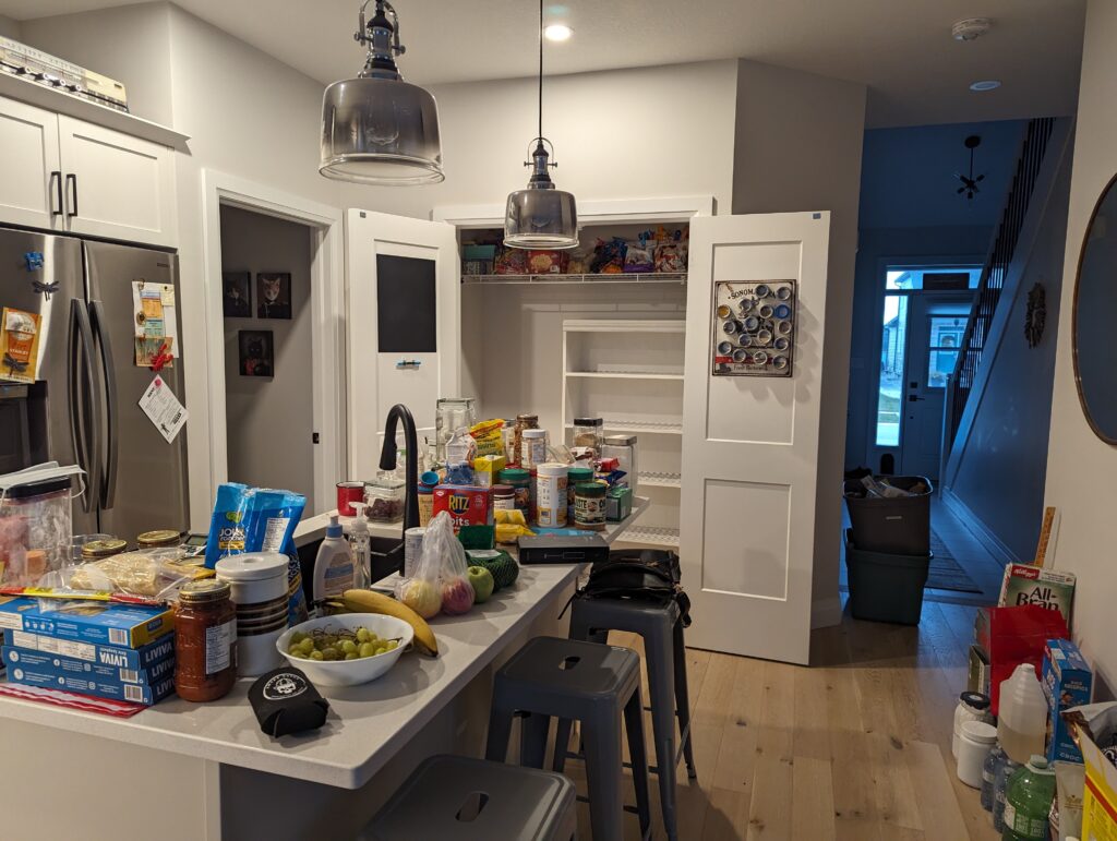 The pantry is cleared out and the bookshelf is empty. Everything from canisters to peanut butter to Ritz crackers can be seen on the island counter. 