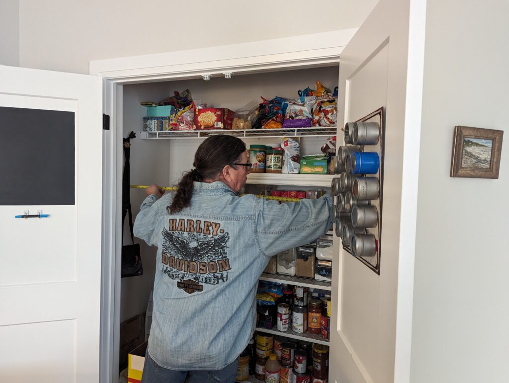 Derek measures the width of the space. He's wearing a denim Harley Davidson shirt and looking serious. 