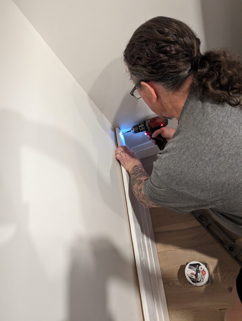 Derek drills a piece of wood into the lower corner of the small room. 