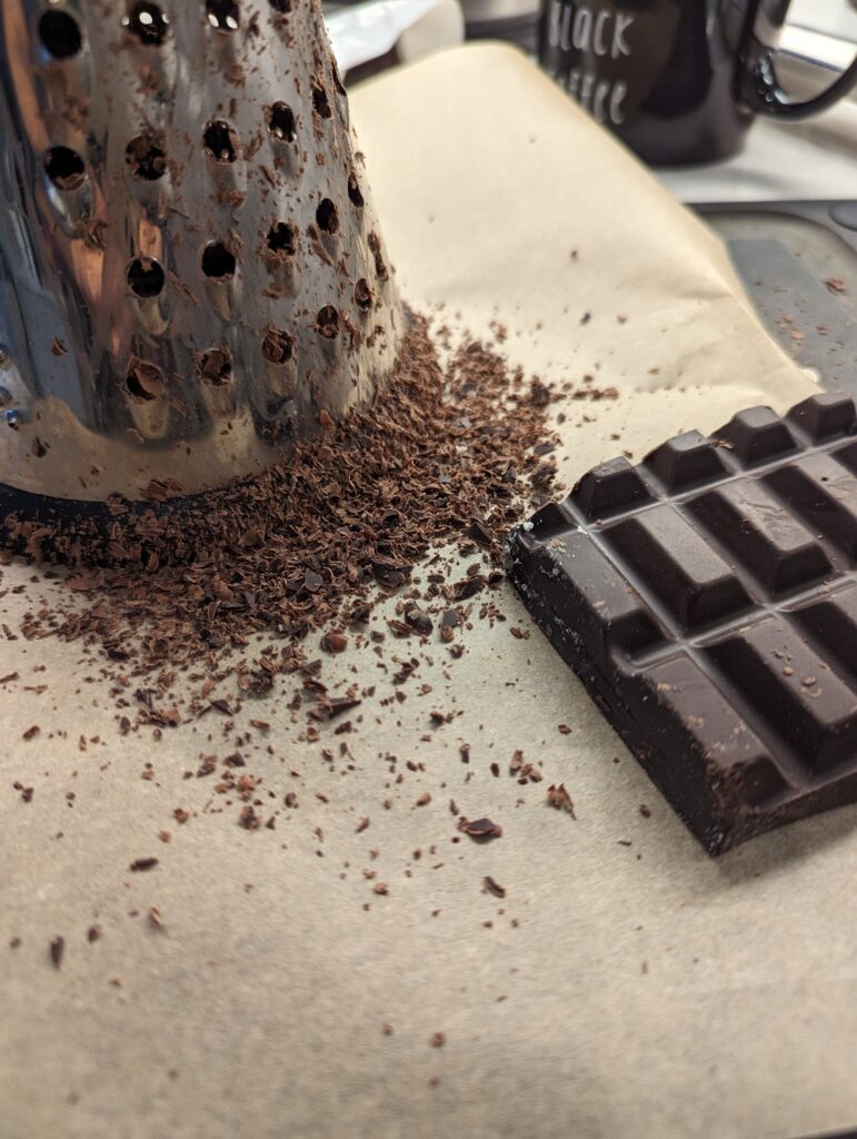 Tall grater on parchment paper. Grated chocolate in the foreground beside a piece of baker's chocolate.