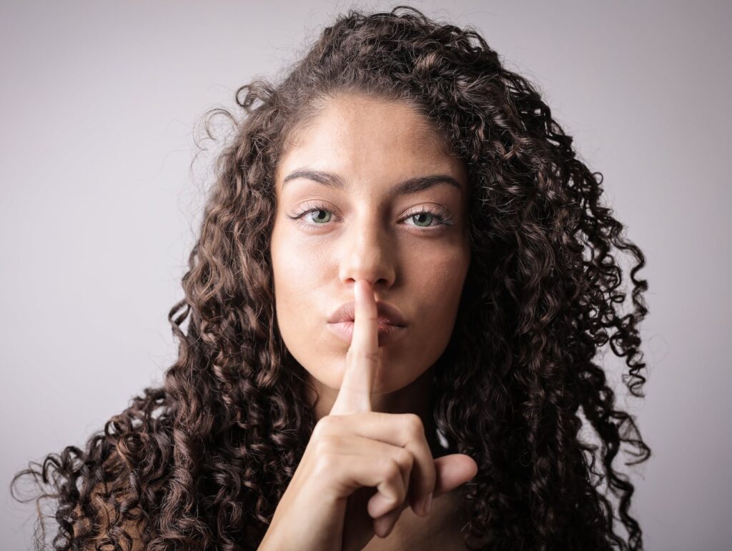 portrait photo of woman with brown curly hair doing the shhh sign