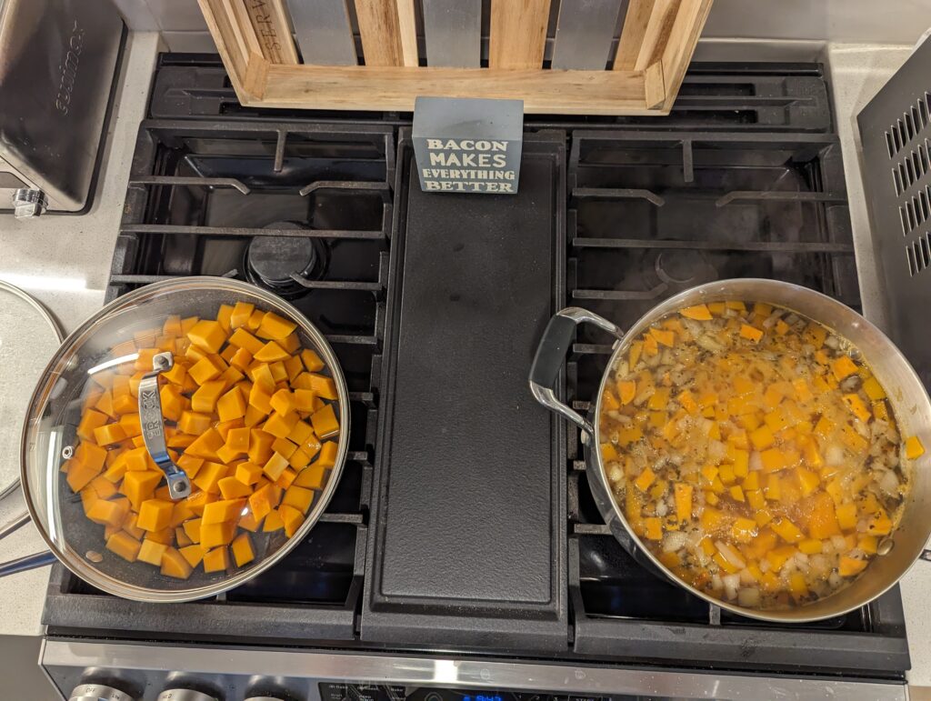 A pan of cubed squash in water on the left of the gas stofe and a pot with cubed squash, sweet potato and onion visible along with spices. 