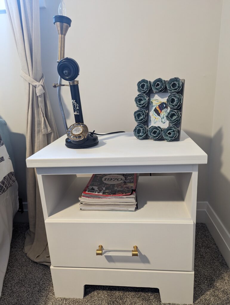 White night table with an open shelf and lower drawer. On top, is an old tall rotary phone whose earpiece is now a lamp. The handle is acrylic with gold tone ends.