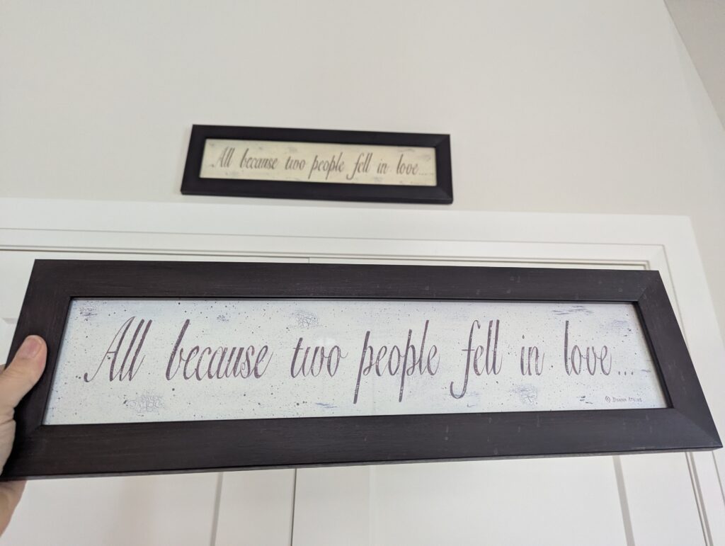 I'm holding a rectangular sign that has script: All because two people fell in love. Above it over the pantry is the same sign except it has a slightly cream-coloured background instead of white. 