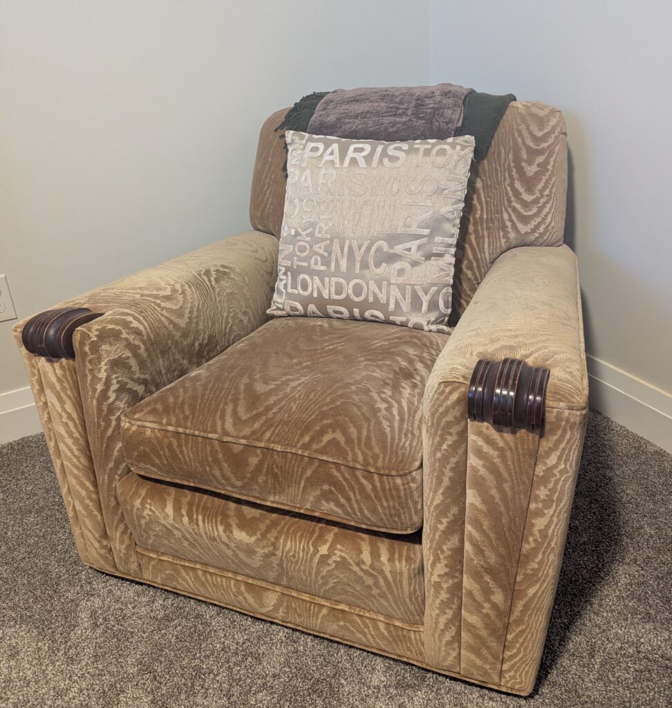 A square upholstered chair with wooden ends on the arms and a neutral cut-velvet upholstery in cream.