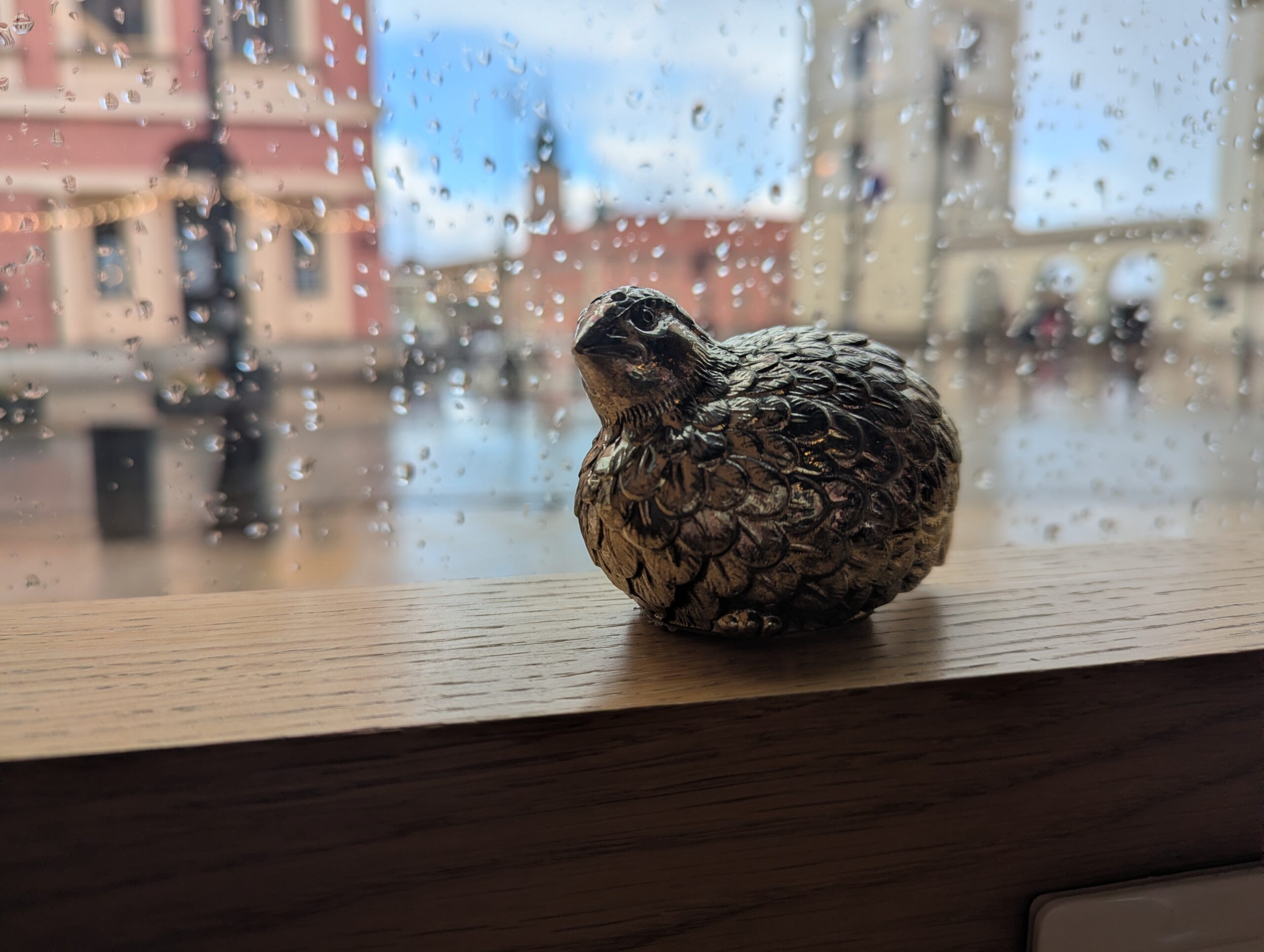 Small, silver bird containing some of my Mom's ashes set on the ledge of a cafe window with raindrops and old buildings in the background