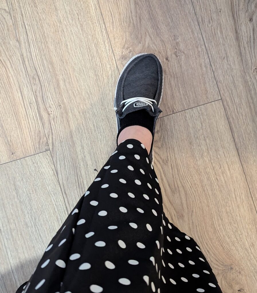 My left food extended to show the dark gray Hey Dude! shoe with a black ankle sock, some bare leg and then the edge of my black and white polkadot skirt.