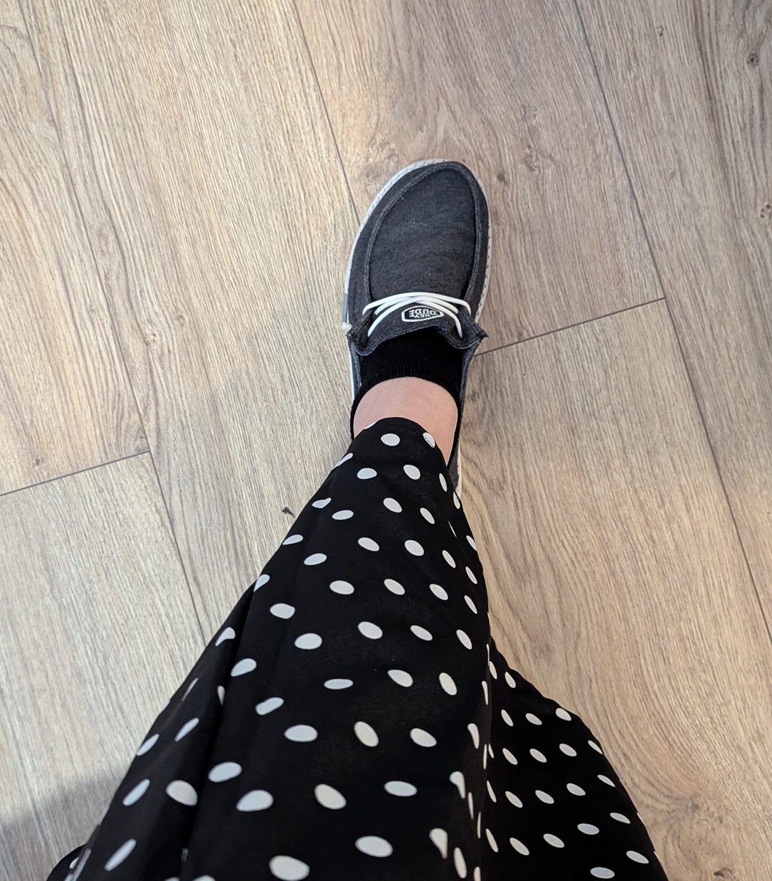 My left food extended to show the dark gray Hey Dude! shoe with a black ankle sock, some bare leg and then the edge of my black and white polkadot skirt.