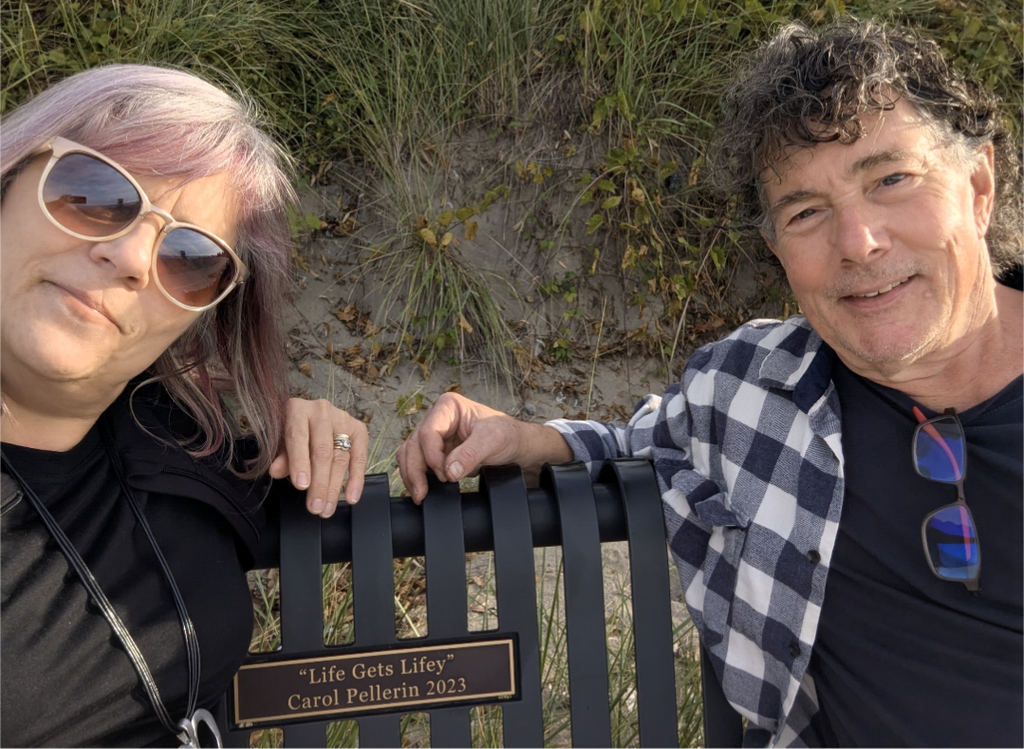 Derek and I seated on a wooden bench on either side of a plaque that reads, Carol Pellerin, 2023, "Life gets lifey."